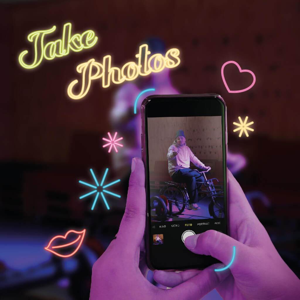 Hand hält ein Smartphone, das eine Person auf einem Fahrrad fotografiert, umgeben von bunten Neonlichtern in Form von Sternen, einem Herz und der Aufschrift "Take Photos" auf einem unscharfen Hintergrund.