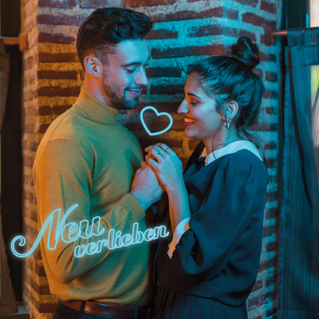 Ein verliebtes Paar, das sich liebevoll anschaut, mit einem neonartigen Schriftzug "Neu verlieben" und einem neonherz über ihnen vor einer Backsteinwand.